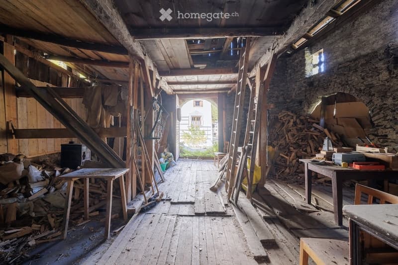 A rustic, unfinished wooden room with exposed beams, wooden floor, and scattered tools and materials. Visible items include a table, stool, chair, ladder, and a pile of wood.