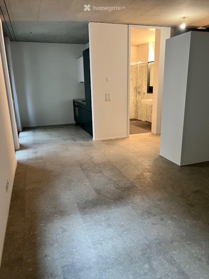 Empty room with tiled floor, white walls, and a small kitchen area visible in the background.