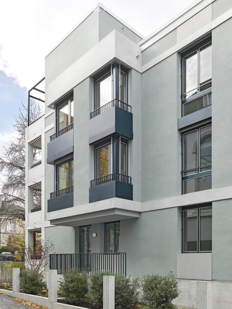 Modern apartment building with 3 floors, gray exterior, balconies on upper floors, entrance with black railings, green bushes and plants surrounding the building