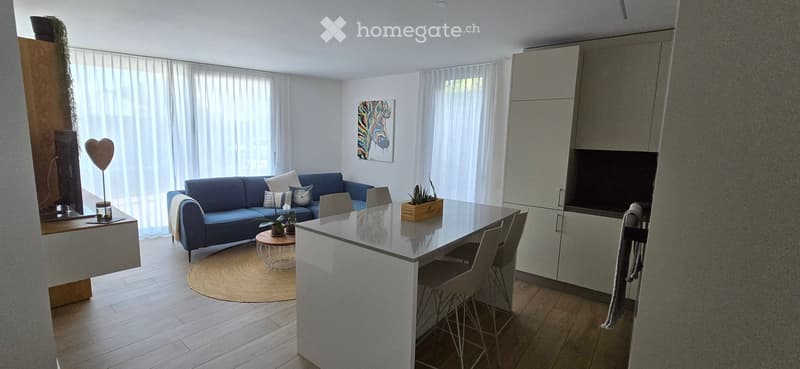 Modern living room with a blue sofa, coffee table, round stool, dining table with chairs, and a cabinet. Large windows with white curtains and a plant on the table.