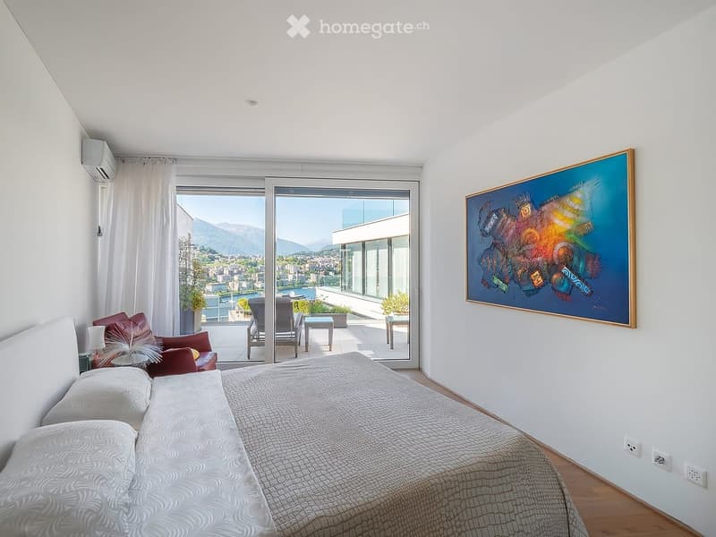 Modern bedroom with large windows offering a scenic mountain view, featuring a bed, a red armchair, a small table, a lamp, and a colorful painting on the wall.