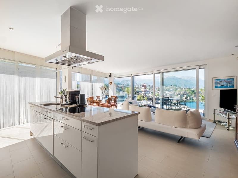 Modern open-plan living and dining area with a white sofa, coffee table, kitchen island, stove, fridge, dining table, chairs, and a television. Large windows offer a scenic view of the city and mountains.