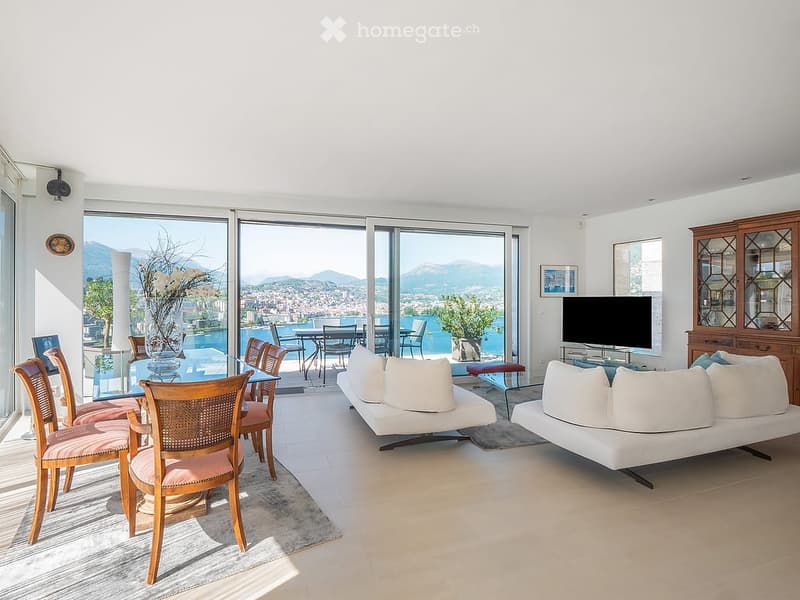 Bright living room with large windows offering a scenic view, furnished with a white sofa, chairs, a dining table, and a television.