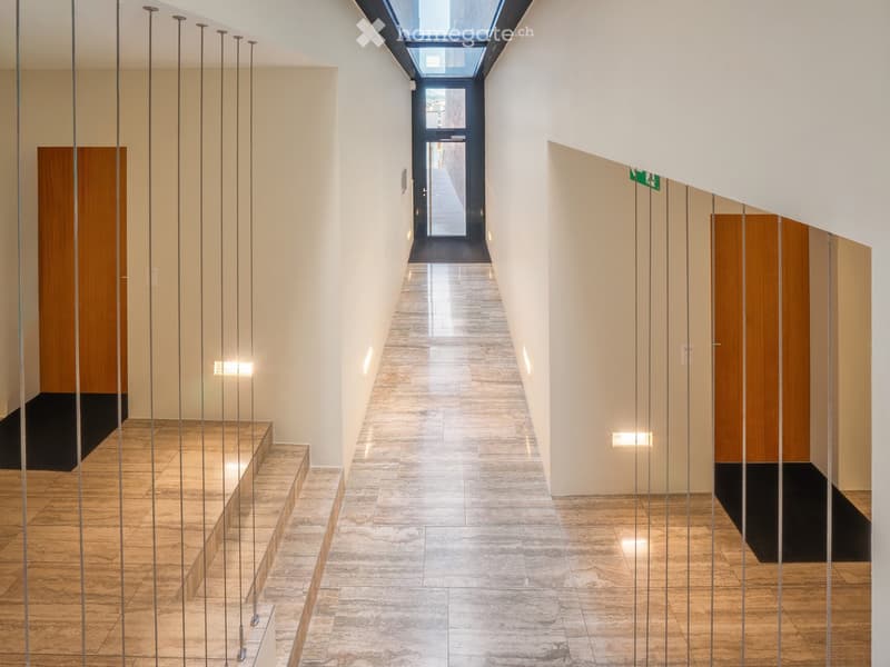 Modern hallway with polished tile flooring, white walls, wooden doors, and a skylight at the far end.