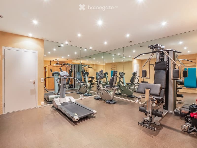 Well-lit fitness room with a variety of exercise equipment including a treadmill, elliptical machine, weight machine, and exercise bike, all reflected in large mirrors.