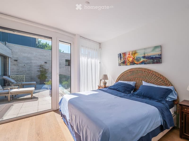 Bright bedroom with a large bed, nightstand, lamp, and a view of a balcony area with outdoor seating.
