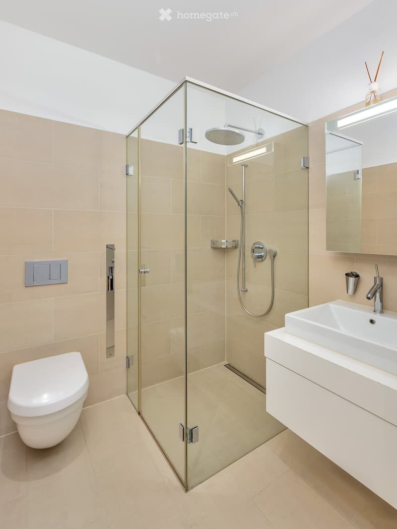 Modern bathroom with beige tiles, glass shower enclosure, toilet, sink, and mirror.