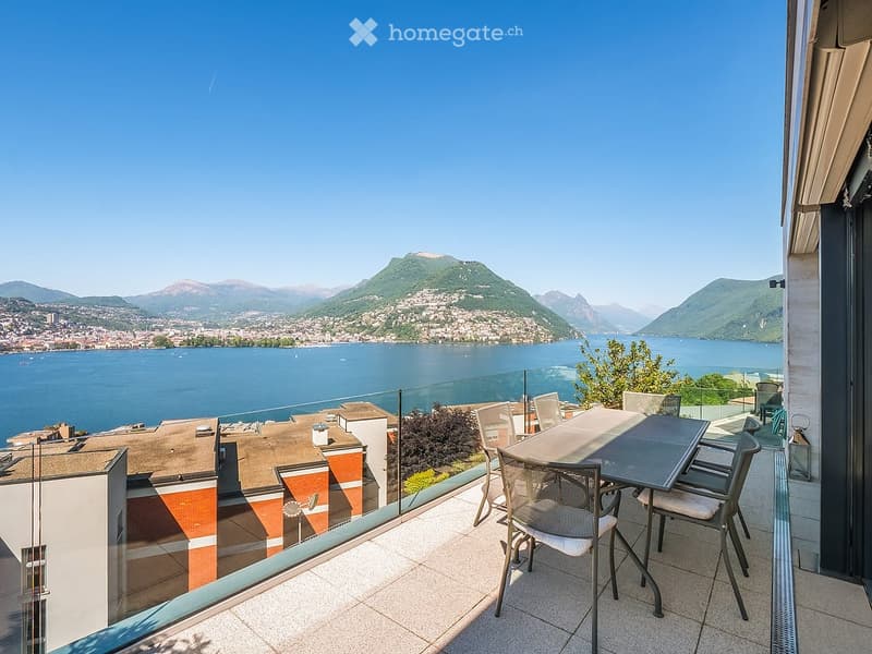 Balcony with a scenic view of a lake and mountains, featuring a table and chairs.