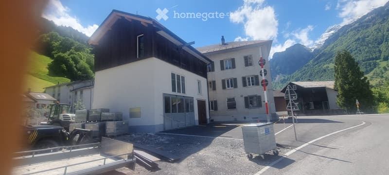 A two-story building with white walls and dark wooden roof accents, situated in a mountainous area with green hills and trees in the background.