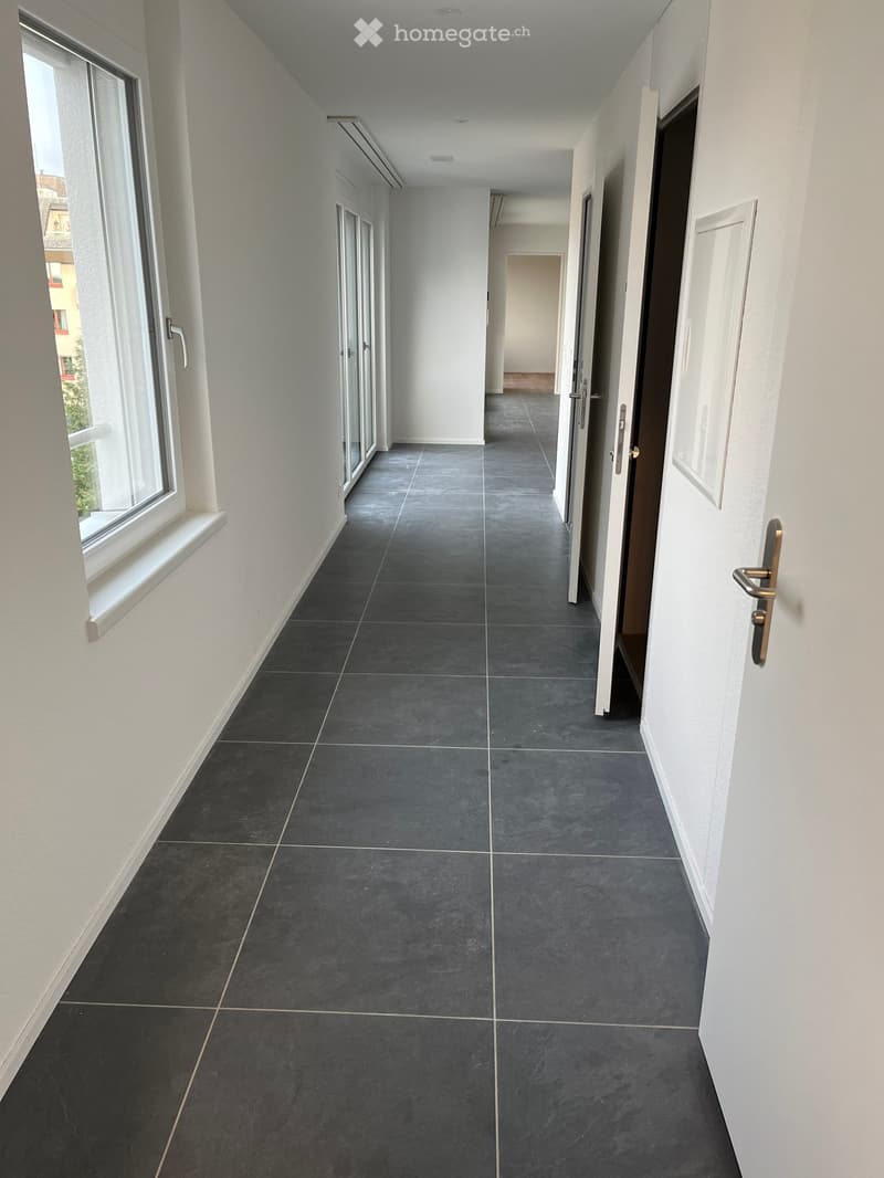 Wide hallway with grey tiled floor, white walls, glass doors and windows, no furniture.
