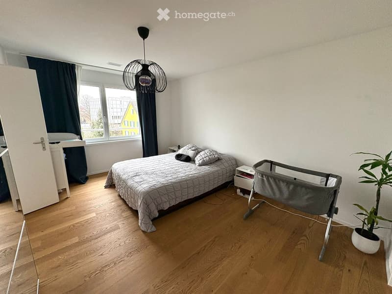 wooden floor, white walls, large window, bed with pillows, bedside table with lamp, hanging lamp, white door, small plant in a white pot, baby crib