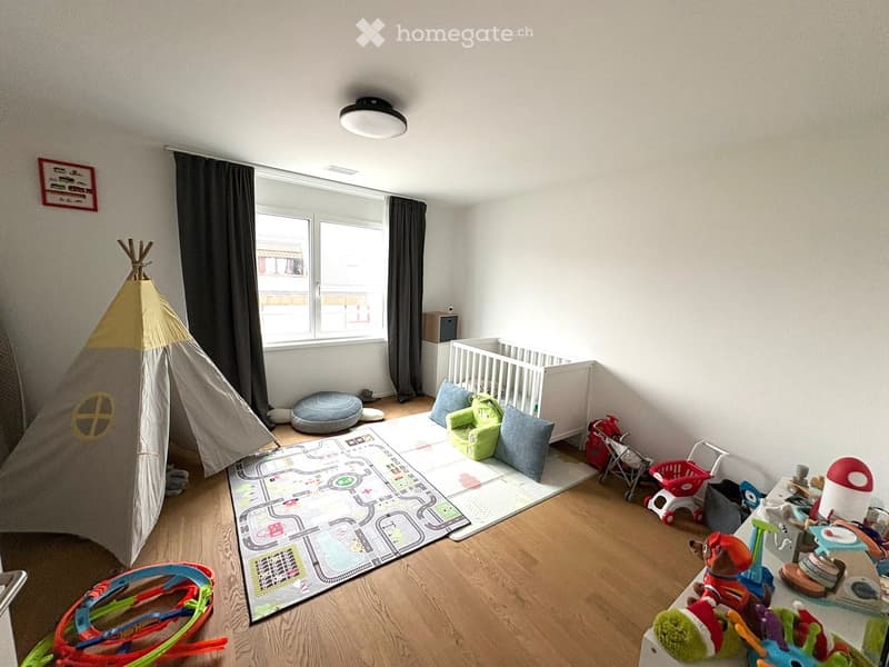 Bright and clean child's room with wooden floor, baby crib, play mat, colorful toys, a tent, white walls, a window with black curtains, and a ceiling lamp.