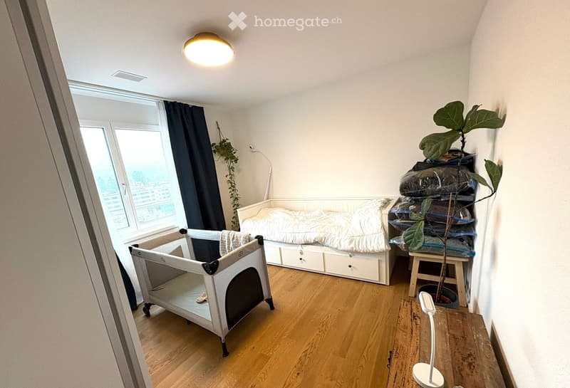 Cozy bedroom with a single bed, nightstand, a small plant, a baby playpen, and a modern lamp. The room features wooden flooring and large windows with dark curtains.
