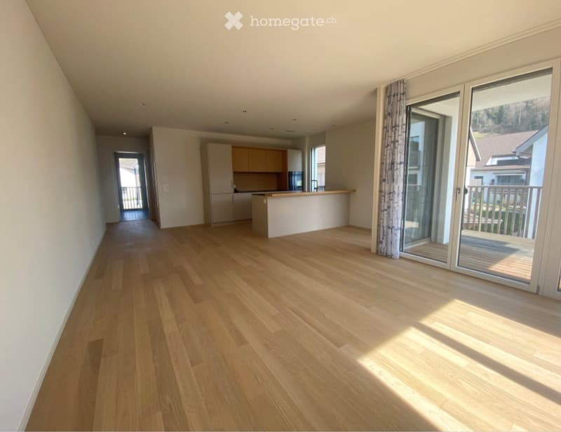 Unfurnished Room. Bright, unfurnished living space with wooden flooring, large sliding glass doors leading to a balcony, and a small kitchen area visible in the background.