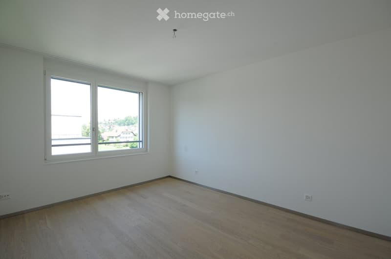 Empty room with light wooden flooring, white walls, and large windows offering a view of the outside area.