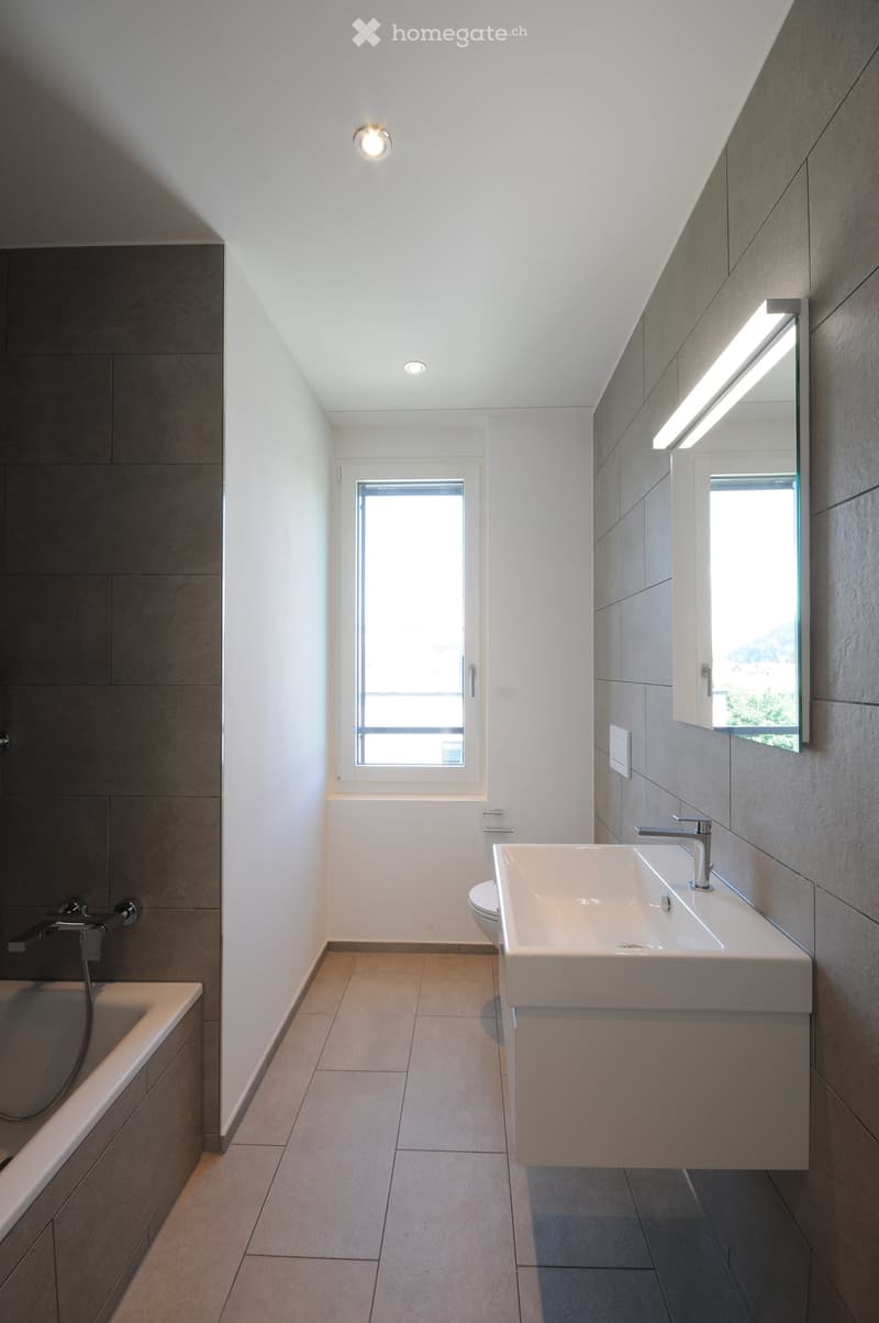Modern bathroom with a white sink, bathtub, toilet, and large mirror. Features recessed lighting and a window providing natural light.