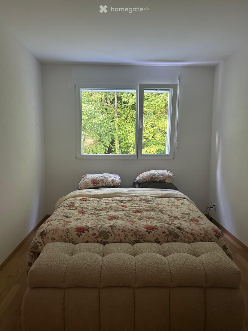 A cozy bedroom with a large bed, floral bedding, pillows, and a tufted bench at the foot of the bed. The room has light-colored walls and wooden flooring, with two windows offering a view of greenery outside.