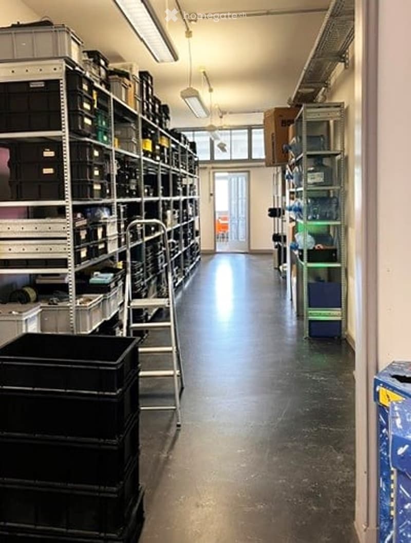 A long, narrow hallway with industrial shelving units on both sides, filled with various storage bins and containers. The floor is polished concrete, and the ceiling has fluorescent lighting. The hallway leads to a door at the far end.