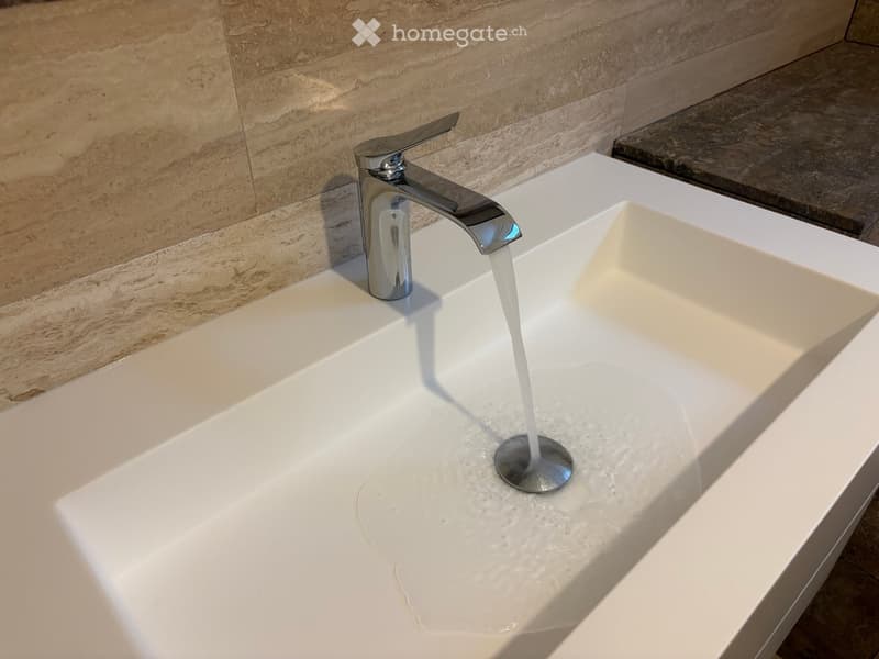 Modern bathroom sink with a sleek, silver faucet and a minimalist white basin, set against a textured beige tiled wall.