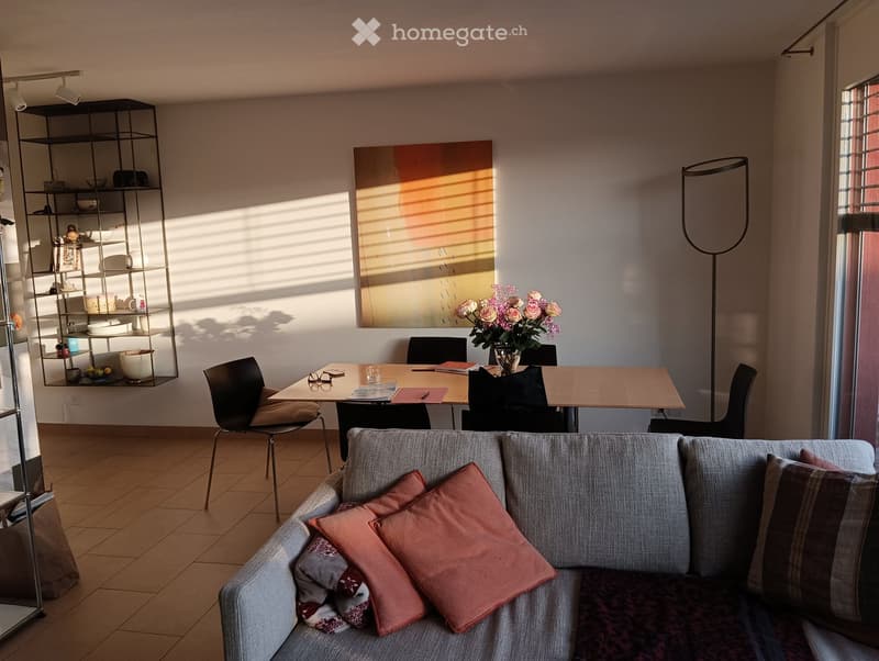 A modern living room with a grey sectional sofa, wooden coffee table, dining table with chairs, and a tall floor lamp. Shelves hold decorative items and books, and a large abstract painting hangs on the wall.