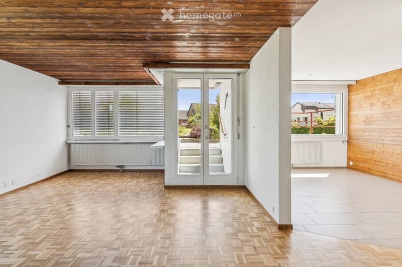Empty room with wooden herringbone flooring, white walls, and a wooden ceiling. Features large windows with blinds, a sliding glass door leading to a staircase, and a radiator under one window.