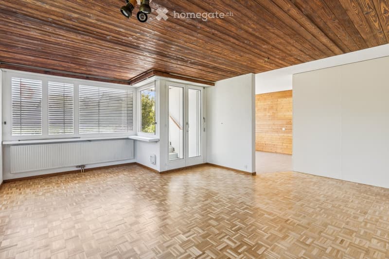 Empty room with wooden herringbone flooring, white walls, large windows with blinds, and a sliding glass door leading to a balcony.