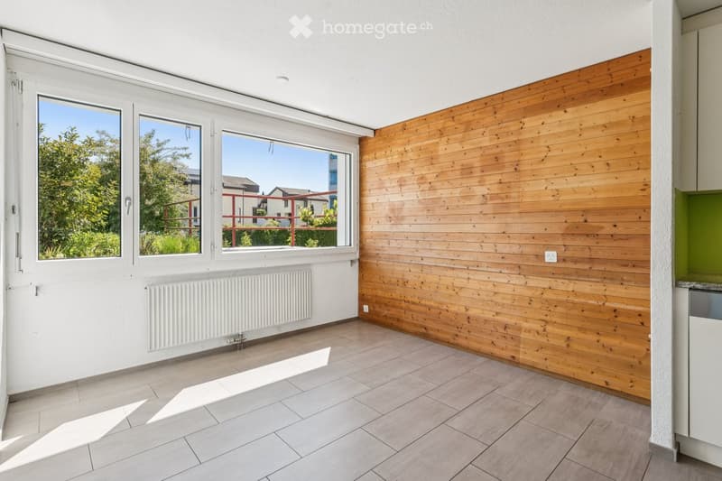 Empty room with tiled floor, white walls, wooden accent wall, large windows, radiator, and a small cabinet.