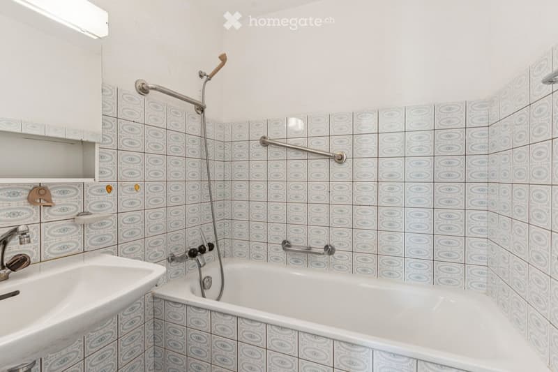 Bathroom with a bathtub, sink, and showerhead. The walls are tiled with a blue and white pattern, and there are handrails for safety.