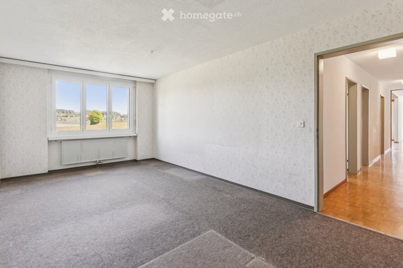 Empty room with grey carpeted floor, white walls, large window with view, radiator, and open doorway leading to hallway.