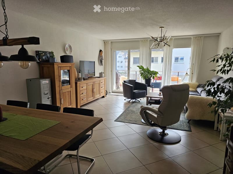 a living room with a wooden table and chairs, an entertainment center, a television, a couch, a recliner, a rug, a lamp, plants, a chandelier, and glass doors leading to a balcony
