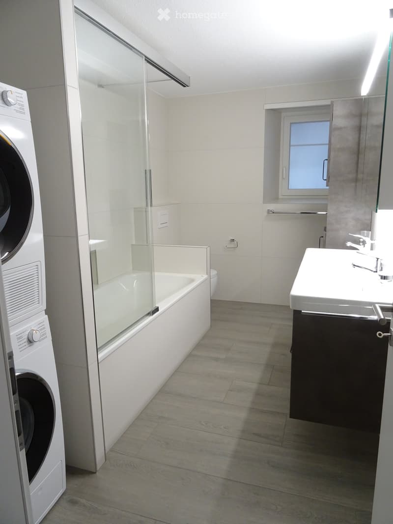Modern bathroom with a bathtub, sink, and washing machine. Features a glass-enclosed shower area and a window providing natural light.