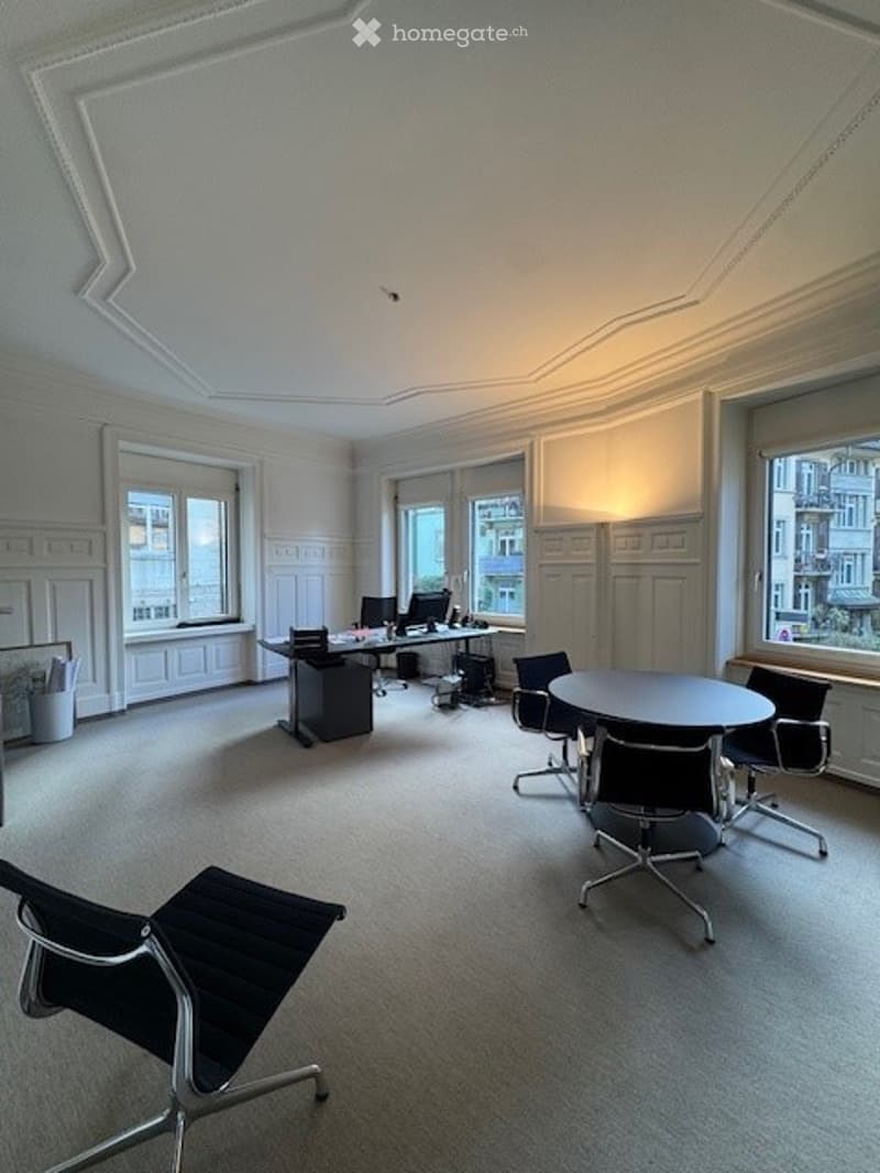 A bright and modern office with white walls, carpeting, and ample windows. It has a desk, computer, and several chairs around a round table. A lamp provides lighting.