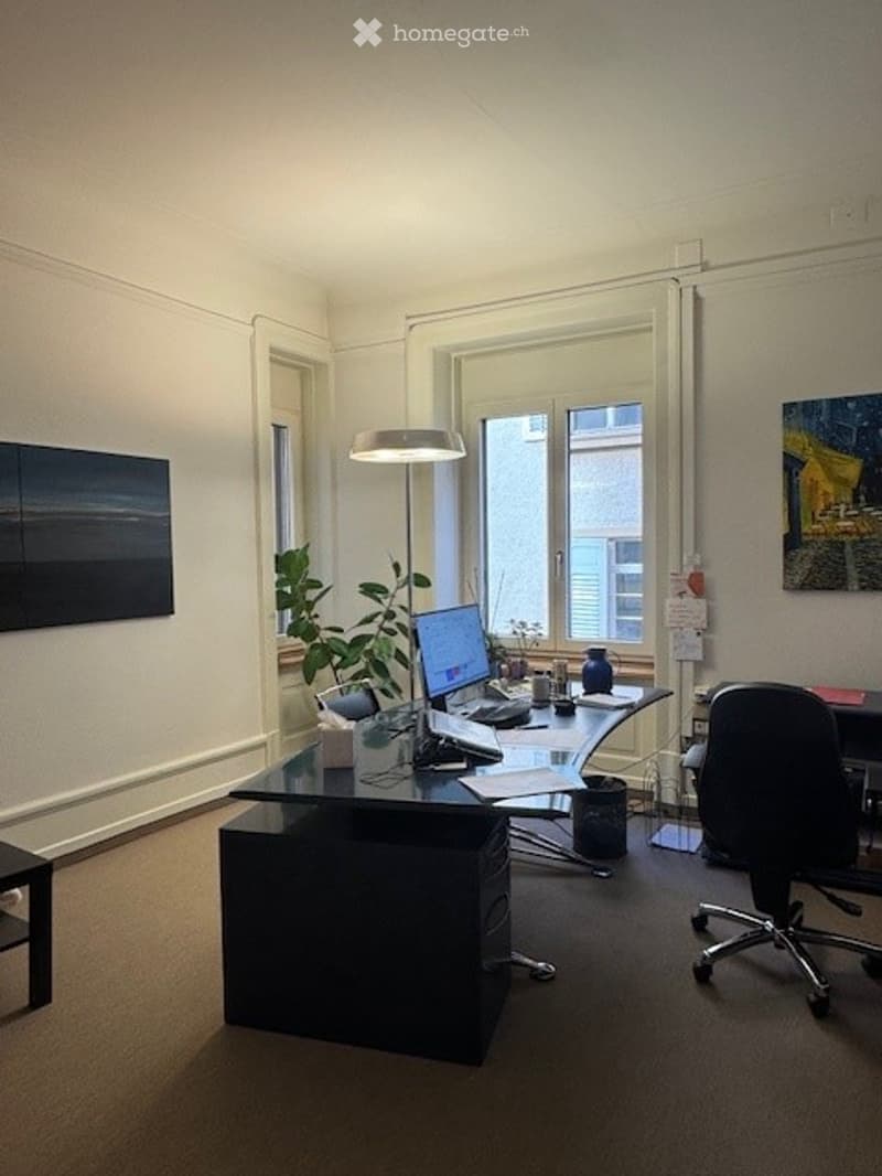 Office room with desk, black swivel chair, computer, plants, lamp, painting, white walls, beige floor.