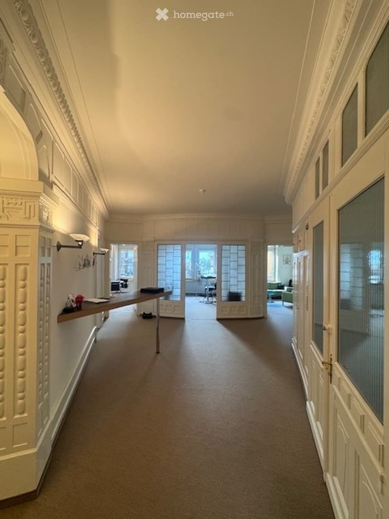 Empty hallway with white walls, beige carpet, glass doors, mounted lamps, shelf, and desk