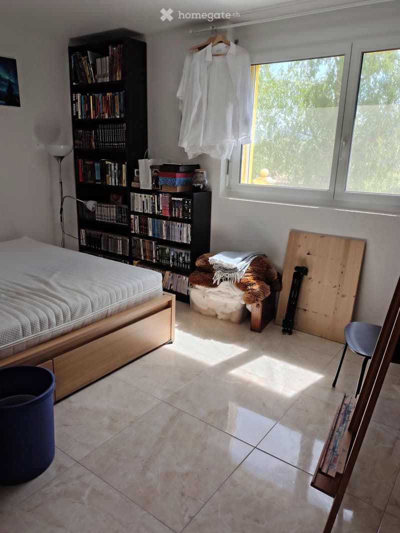 Bright bedroom with a wooden bed, large bookshelf, a small stool with a blanket and teddy bear, a chair, and a lamp. Natural light streams in from a large window.