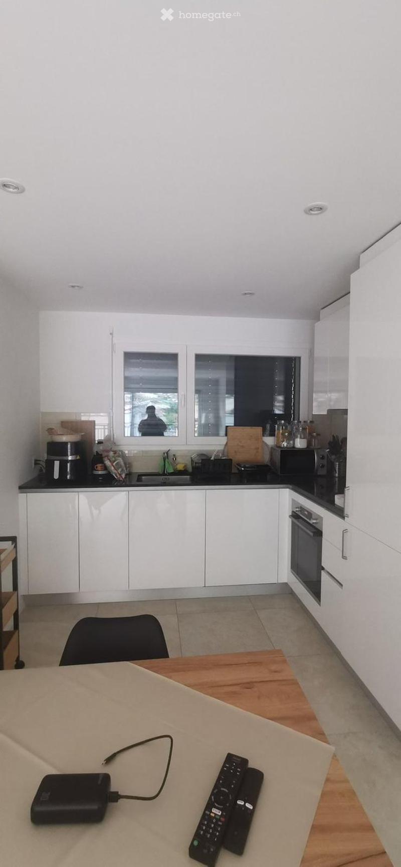 Modern kitchen with white cabinets, black countertops, and a wooden dining table with a chair. Visible appliances include an oven, microwave, and coffee maker. A remote control and power bank are placed on the table.