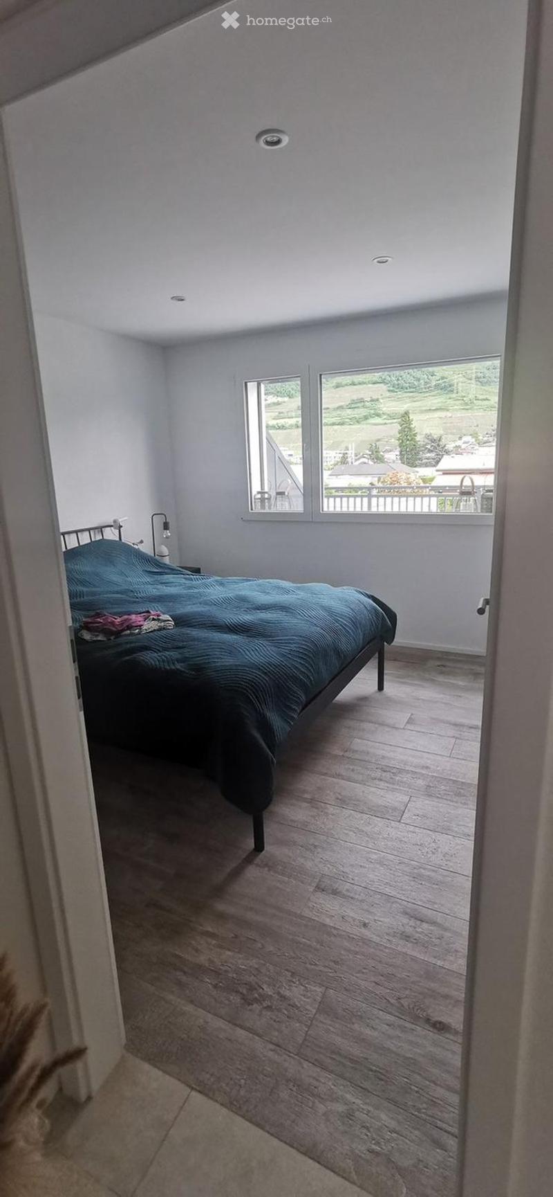 Bright bedroom with wooden flooring, large windows offering a scenic view, a neatly made bed with a blue duvet, and a nightstand with a lamp.