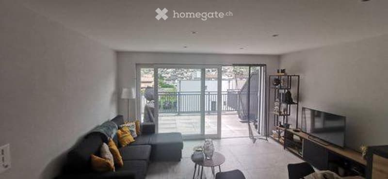 Modern living room with large sliding glass doors leading to a balcony, grey sectional sofa with orange throw pillows, coffee table, floor lamp, TV stand, and a small shelf with decorative items.