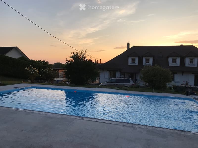 A swimming pool in front of a house during sunset, with surrounding greenery and a parked car.