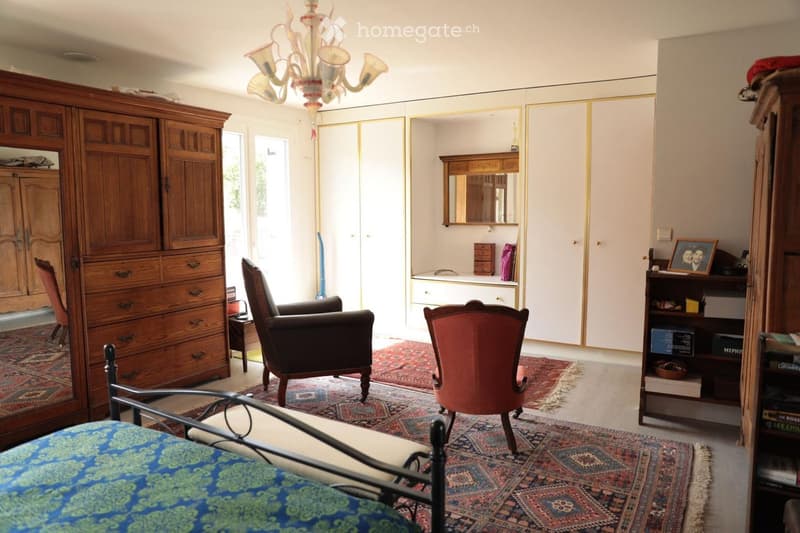 Spacious bedroom with a large wooden wardrobe, a bed with a patterned blue bedspread, two armchairs, a small table, a mirror, and a chandelier.