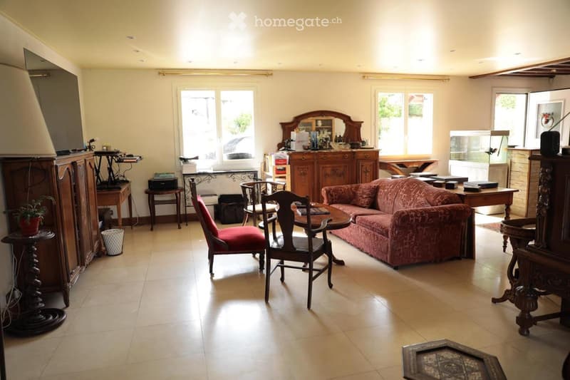 Bright living room with beige walls, tiled floor, large windows, and natural light. Features a red patterned sofa, wooden chairs, a small round table, a wooden cabinet, a mirror, and a lamp.