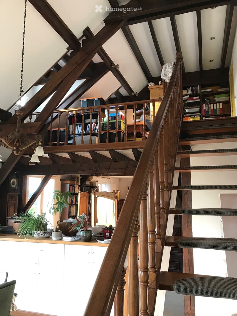 Wooden staircase with a wooden railing and a chandelier, leading to a loft area with bookshelves and storage boxes. The lower level features a white cabinet, potted plants, and a window letting in natural light.