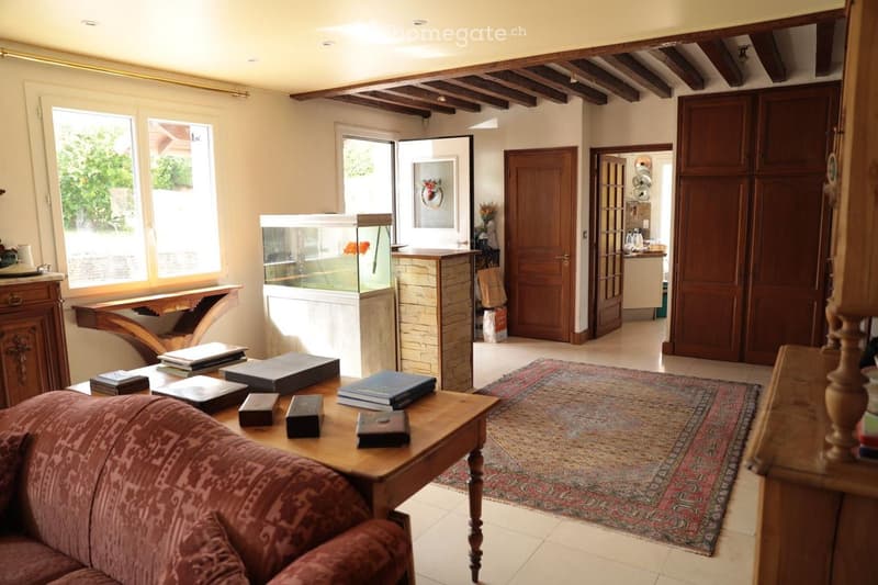 Living room with a red sofa, wooden coffee table with books, fish tank, and a patterned rug on the floor. The room features wooden beams on the ceiling and large windows letting in natural light.