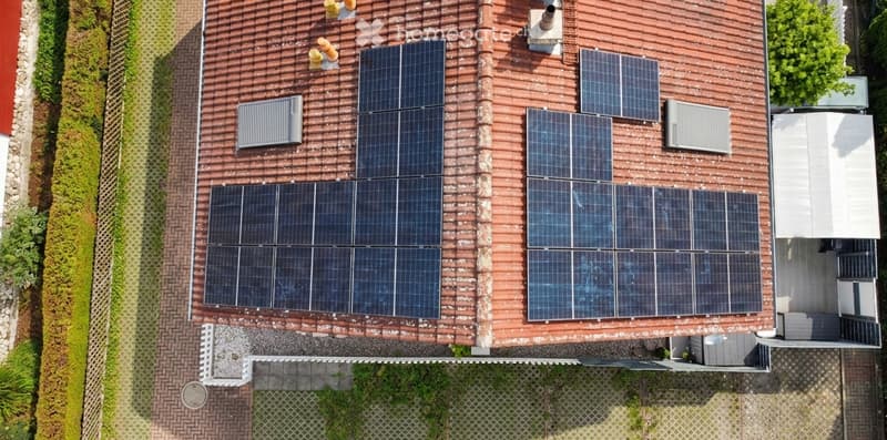 Top-down view of a house roof covered with multiple solar panels, surrounded by a brick-paved area and greenery.