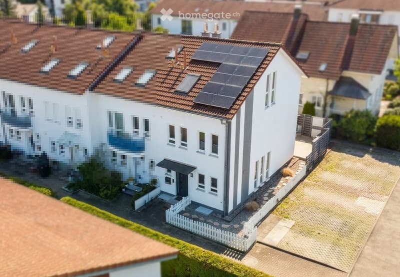 A modern two-story house with a white exterior, brown tiled roof, multiple windows, and solar panels on the roof. The house features a small front yard with a white picket fence and a paved driveway.