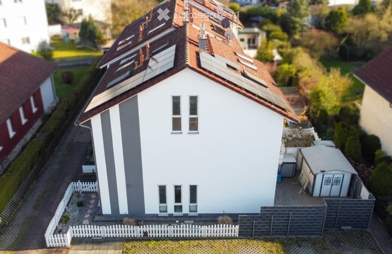 A modern two-story house with white walls, grey accents, and a red-tiled roof. The house features multiple windows, solar panels on the roof, and a small garden area enclosed by a white picket fence.