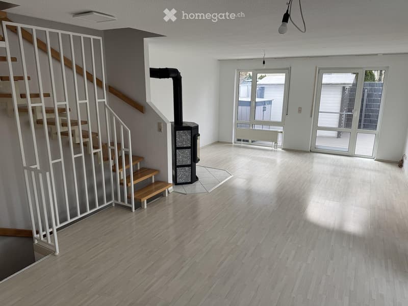 Empty room with light wooden flooring, white walls, a staircase with wooden steps and white railings, and a black wood-burning stove in the corner.