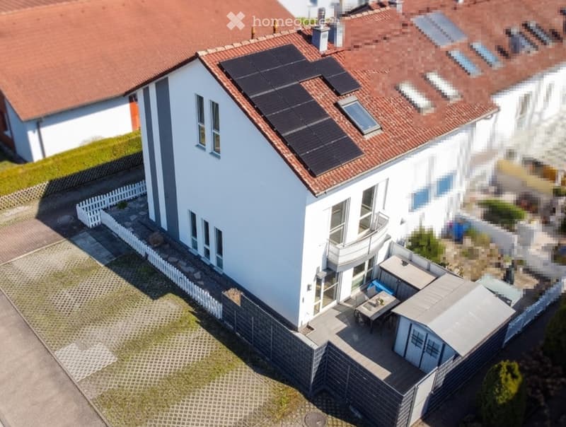 A modern two-story house with white walls, red-tiled roof, solar panels, and a small patio area with outdoor furniture.