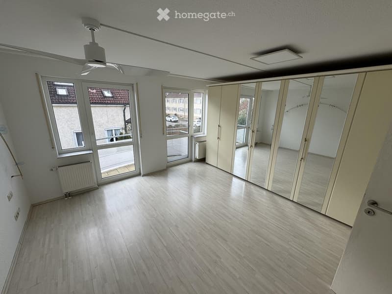 Empty room with wooden flooring, large windows, and a sliding door leading to a balcony. The room has white walls and a ceiling fan.