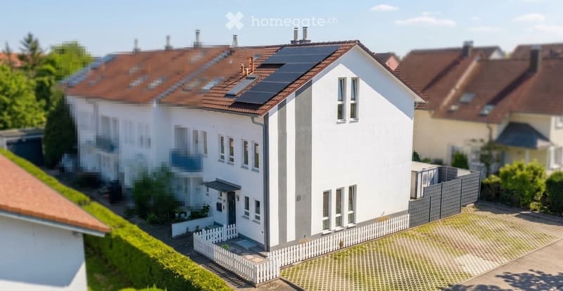 Modern two-story house with white exterior, solar panels on the roof, multiple windows, and a small garden area with a white picket fence.